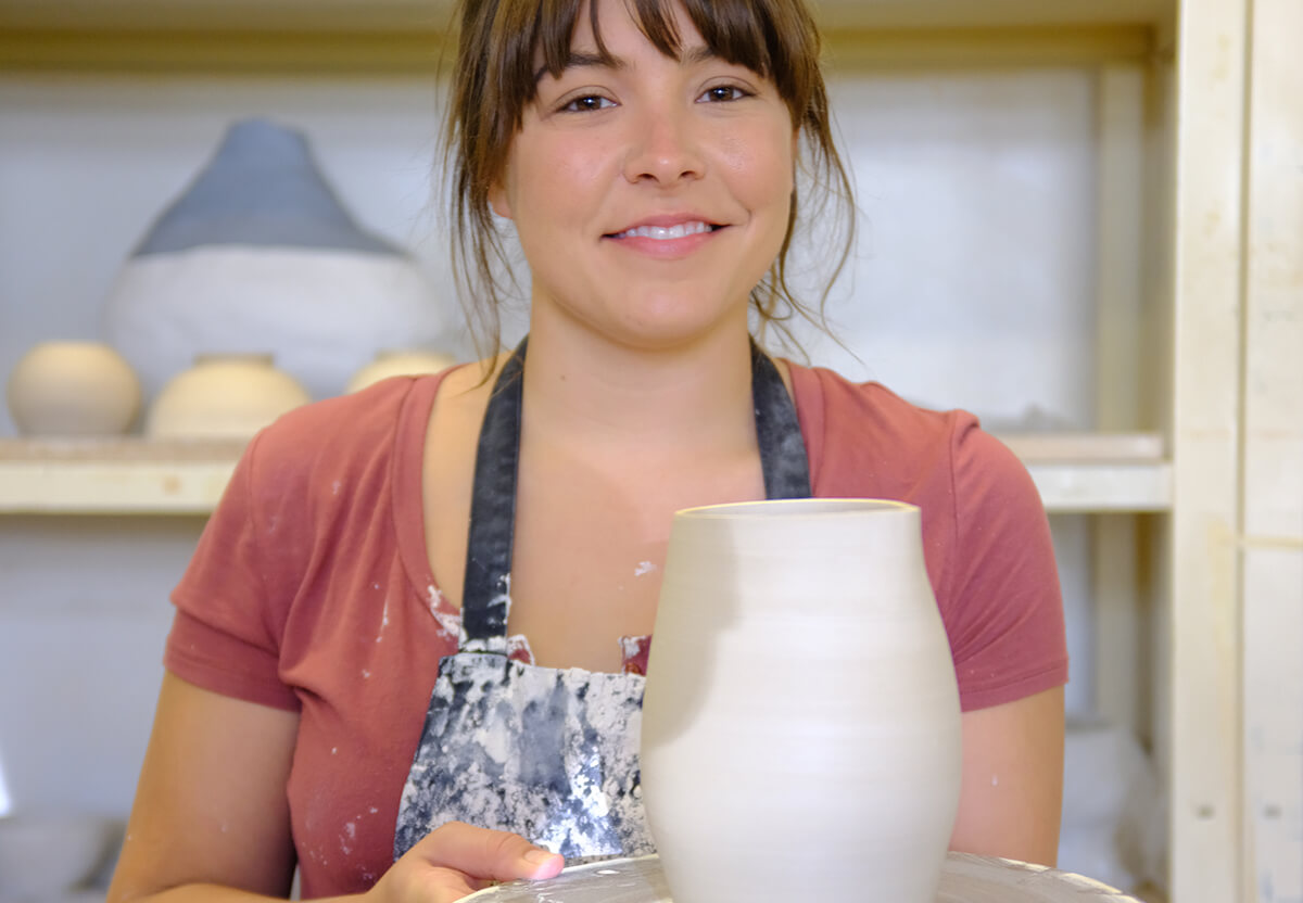 Photo of a ceramics student holding a pot on a stand in studio on campus