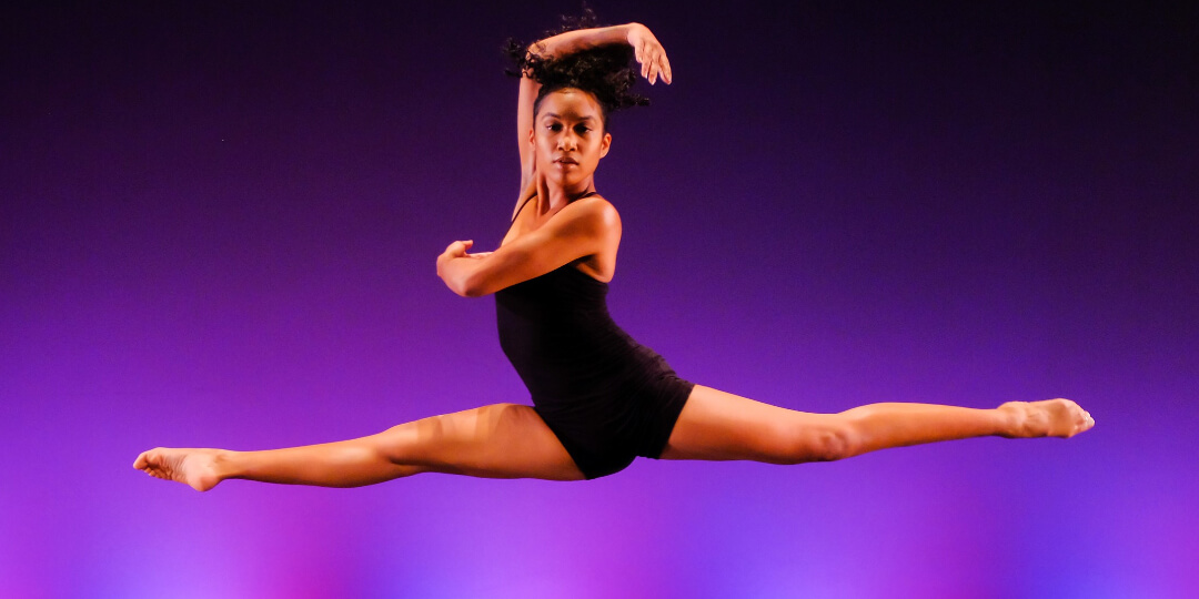 Dancer in mid-air with legs split, left arm across her chest and right arm arched around her head