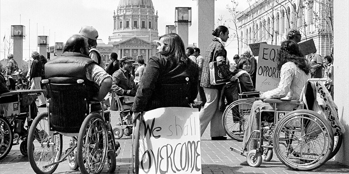 Protesters in wheelchairs by San Francisco City Hall with one holding sign with text We Shall Overcome