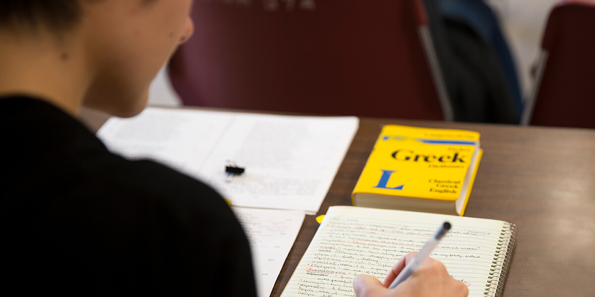 Photo of student writing in notebook with Greek dictionary on her desk