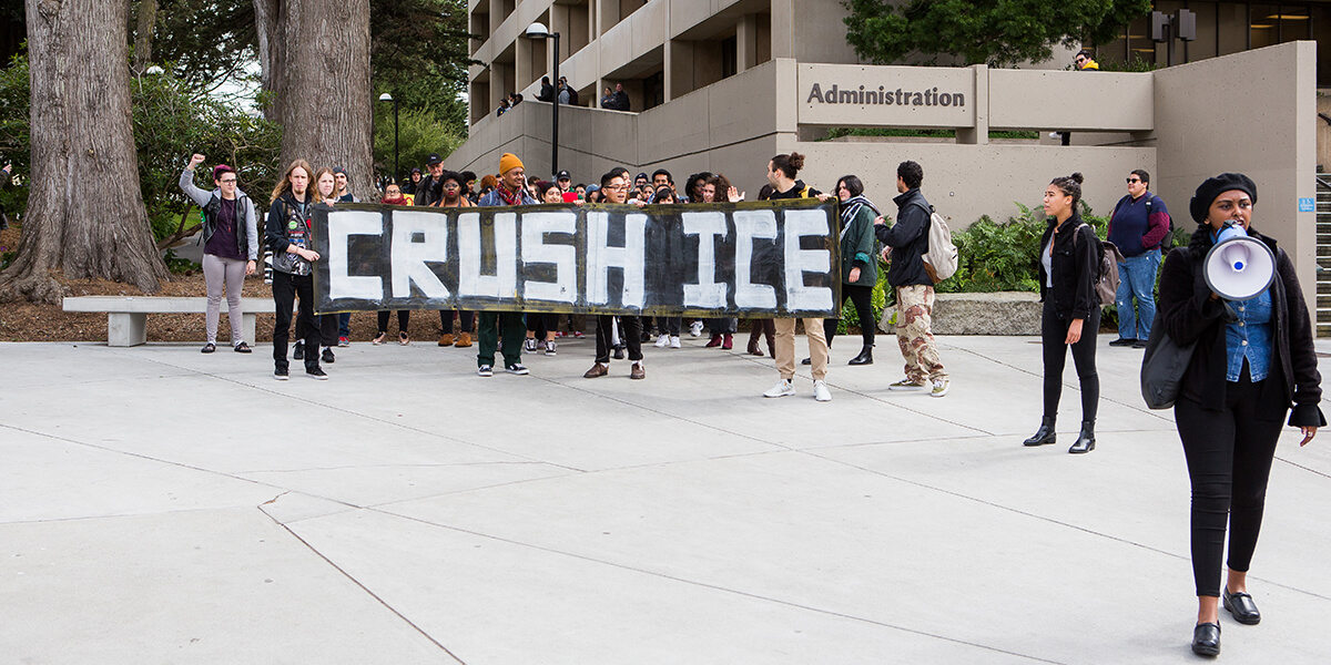 Photo of a 2018 anti-deportation protest at SF State with students holding sign that reads Crush ICE
