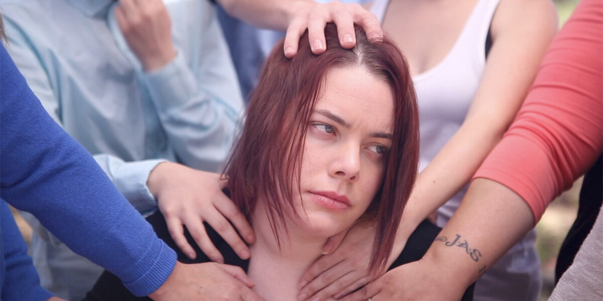 A young woman looks off screen as people’s hands touch her face and upper body. The people’s faces are not in frame.