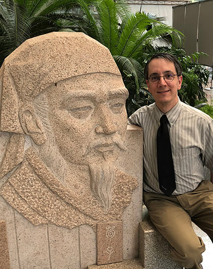 Photo of Eric Hutton seated next to a Confucius statue