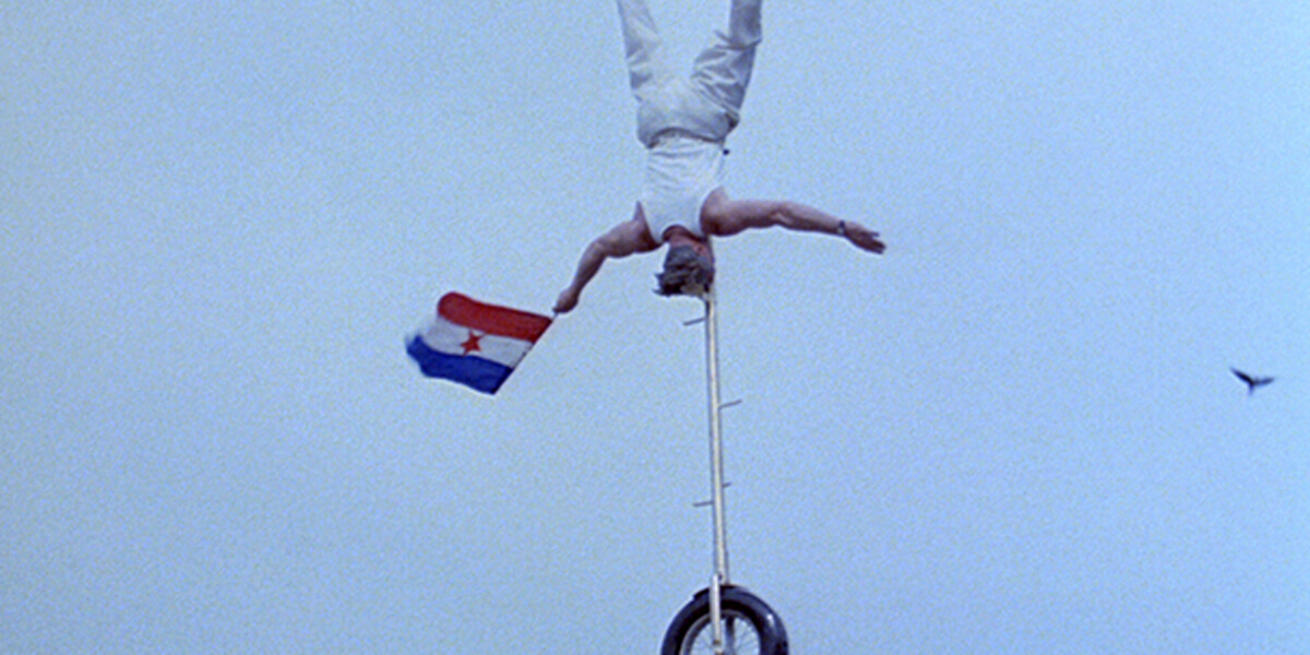 Still of man balancing on a unicycle on his head