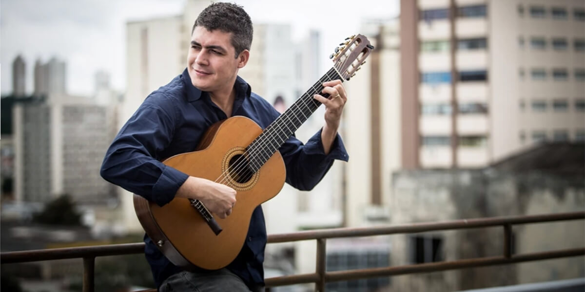 Photo of Julio Lemos playing guitar outdoors on a rooftop near high-rise buildings