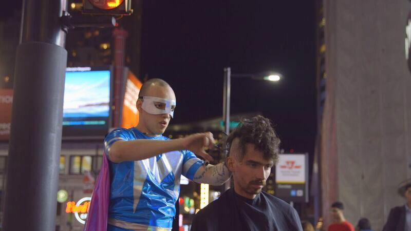 Photo of man in superhero costume cutting a young man's hair outdoors
