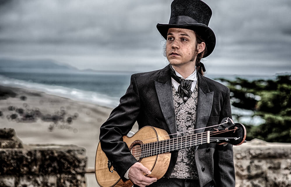 Photo of Jon Mendle dressed in suit and top hat holding guitar at a beach