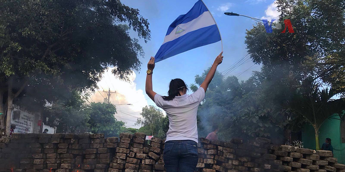 Photo of a woman standing near a burning barricade holding the national flag of Nicaragua