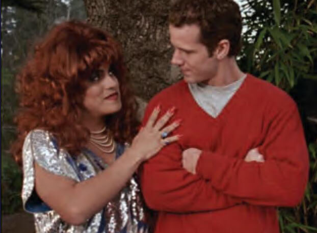 Person in drag embraces a man in a red V-neck sweater who is folding his arms