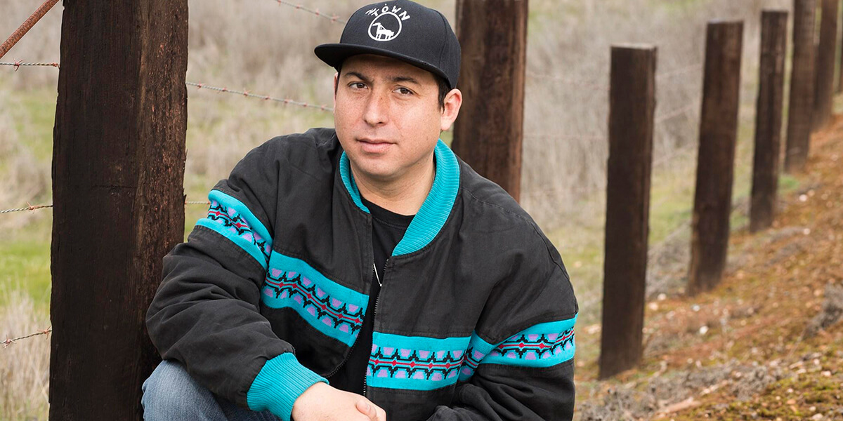 Photo of Tommy Orange kneeling in front of a fence