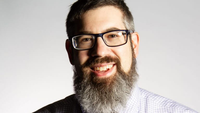 Photo of Jordan Rosenblum smiling with a long graying beard and wearing eyeglasses