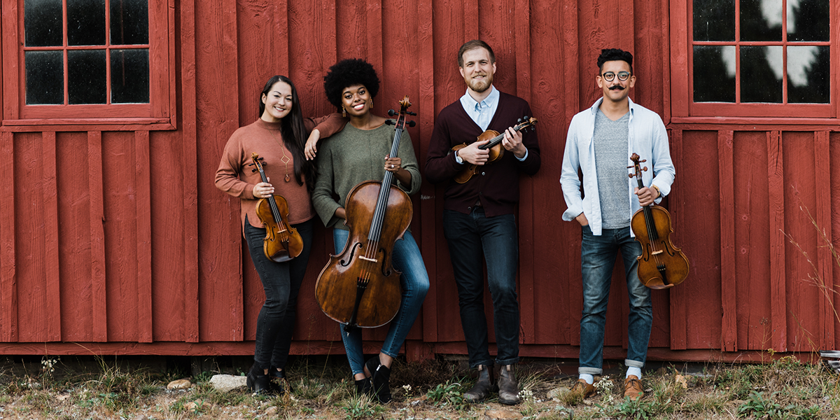 Photo of Thalea String Quartet holding string instruments and standing in front of a barn