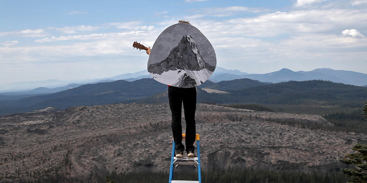 Still of man standing on ladder on mountain holding guitar