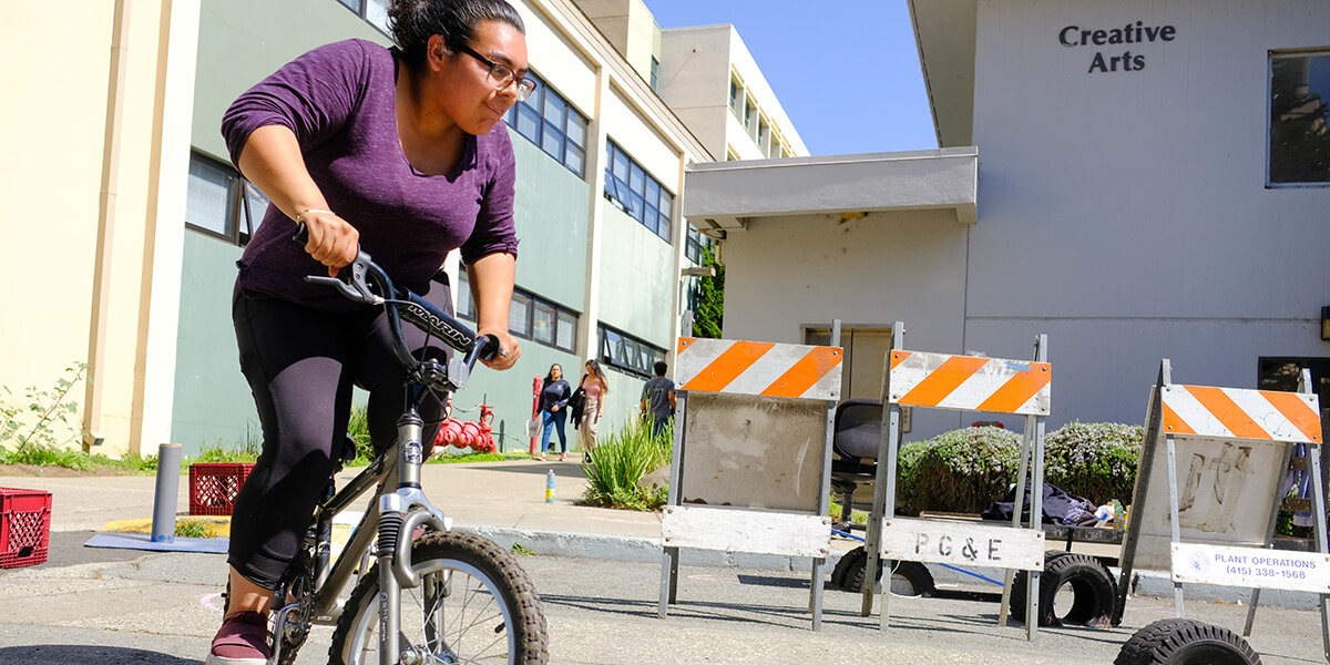 Student Cindy Magana riding a bicycle through an obstacle course on campus