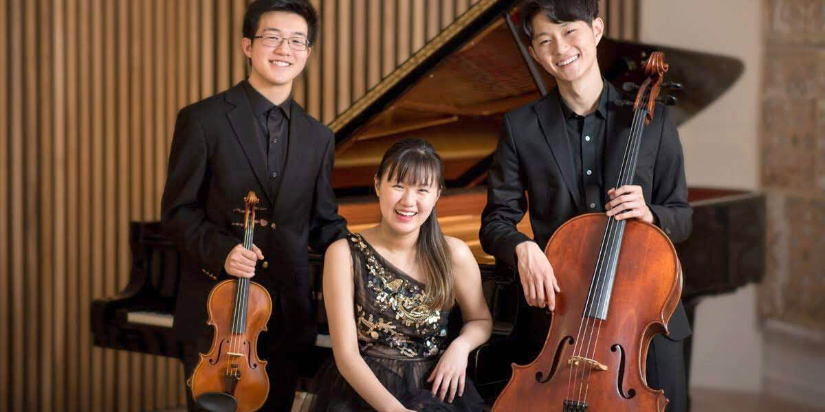 Photo of the Bach Trio holding their instruments in front of a piano