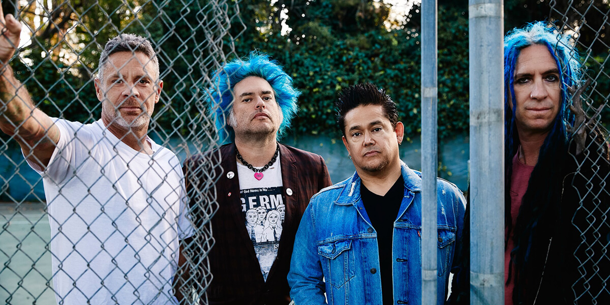 The four members of NOFX standing near a broken chain-link fence at a park