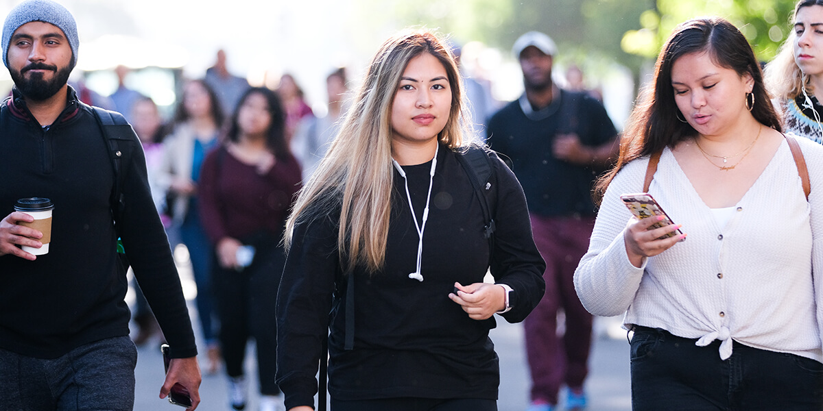 Photo of students walking on campus while holding cell phone and coffee cup