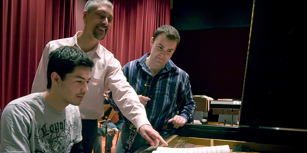 Photo of Cyrus Ginwala and two students looking at sheet music, which is on a piano