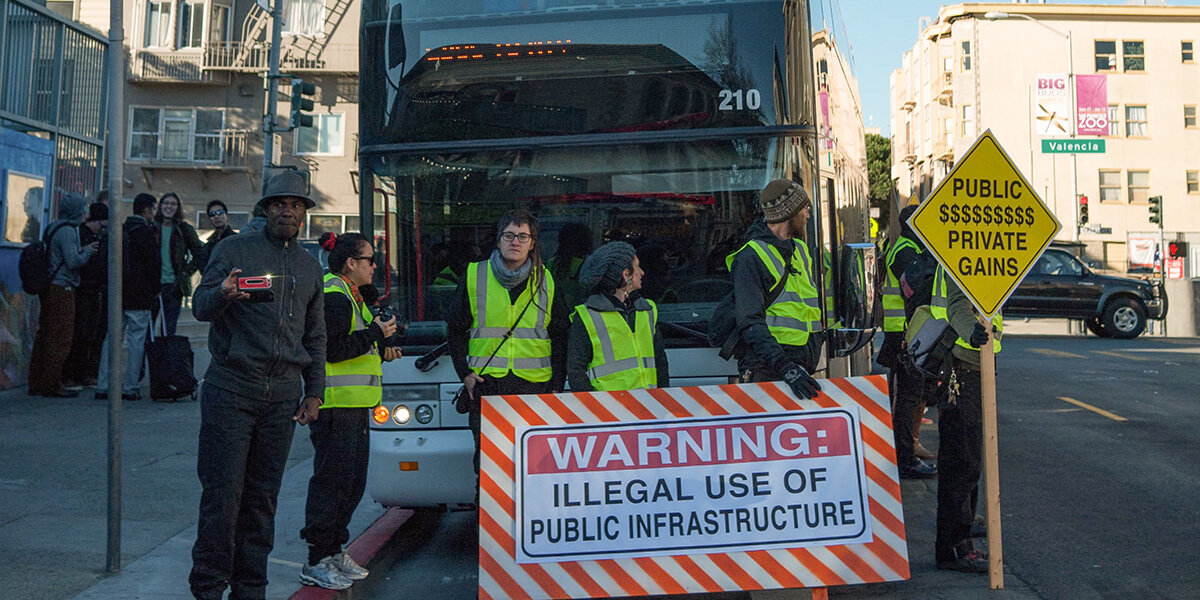 Photo of protestors blocking a private technology-industry commuter bus holding sign Warning: Illegal use of Public Infrastructure