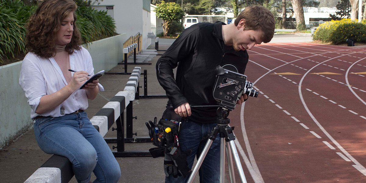 By the Cox Stadium track, a student works with an celluloid film camera while another takes notes
