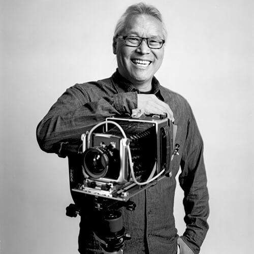 Black and white photo of Paul Kitagaki resting his right arm on a large camera
