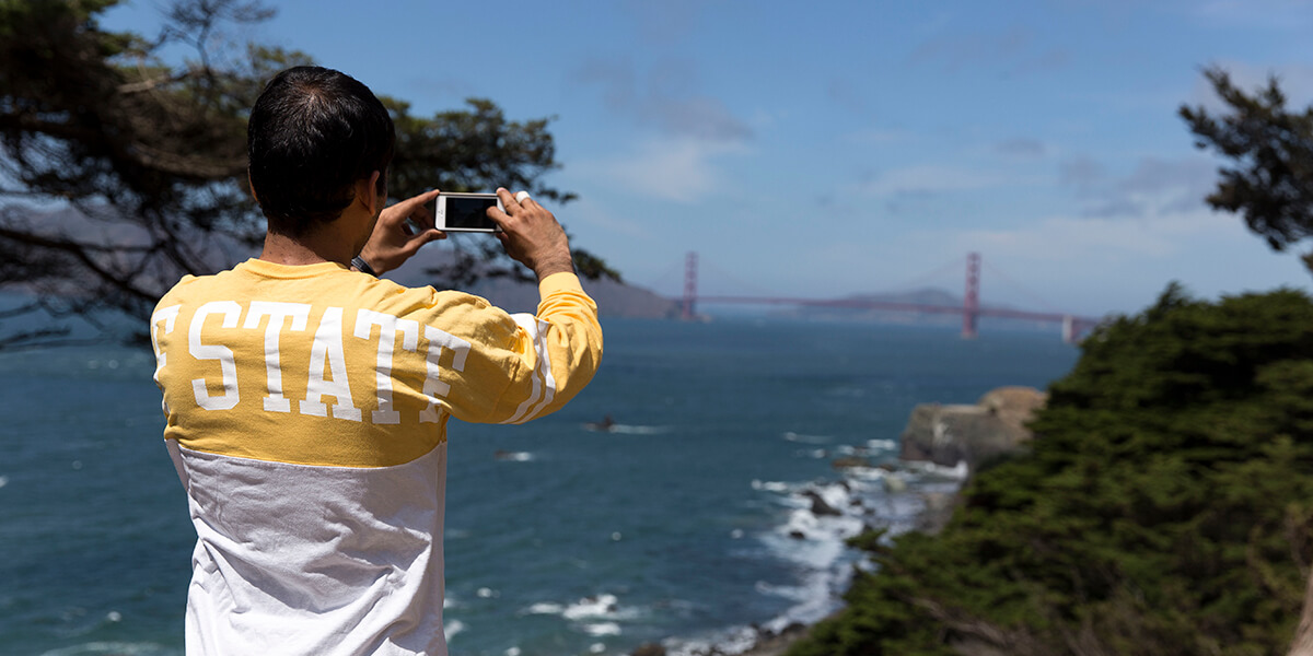 A person wearing an SF State long-sleeve T-shirt uses a cell phone to take a picture of the Golden Gate Bridge