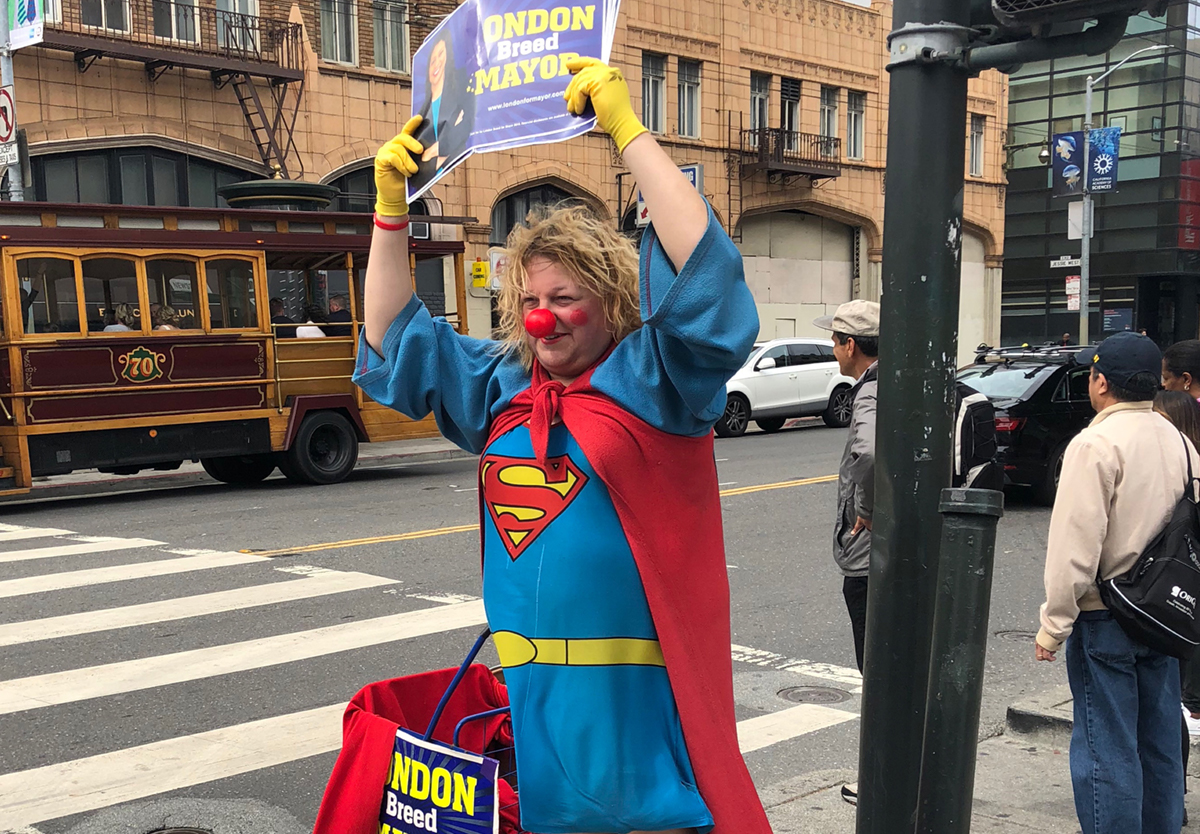 Photo of man in Superman costume and clown nose on street corner holding London Breed for Mayor sign