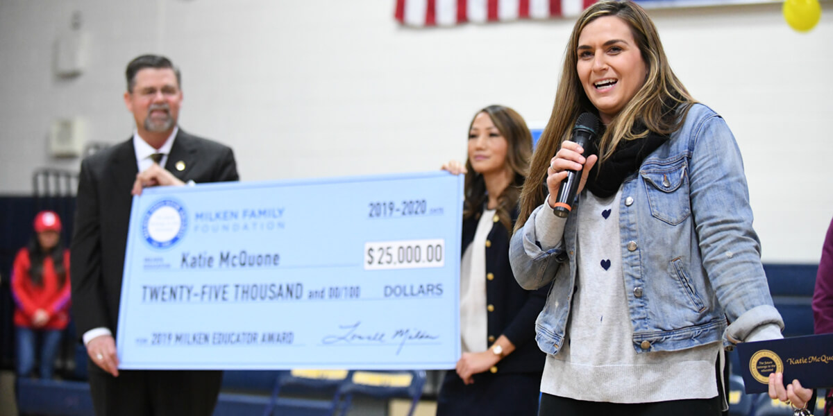 Photo of Katie McQuone speaking with microphone while two other people hold up large check