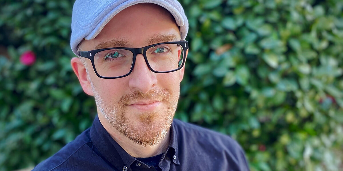 Photo of Joshua McVeigh-Schultz standing in front of a bush wearing eyeglasses, blue shirt and gray hat