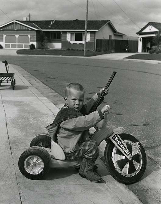 Black and white photo of young boy riding tricycle toting a toy gun