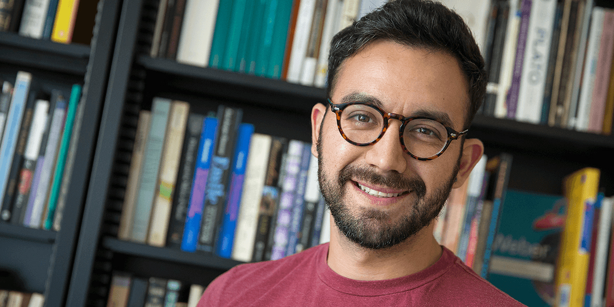 Photo of David Peña-Guzman smiling in front of a bookshelf wearing eyeglasses and wine-colored T-shirt