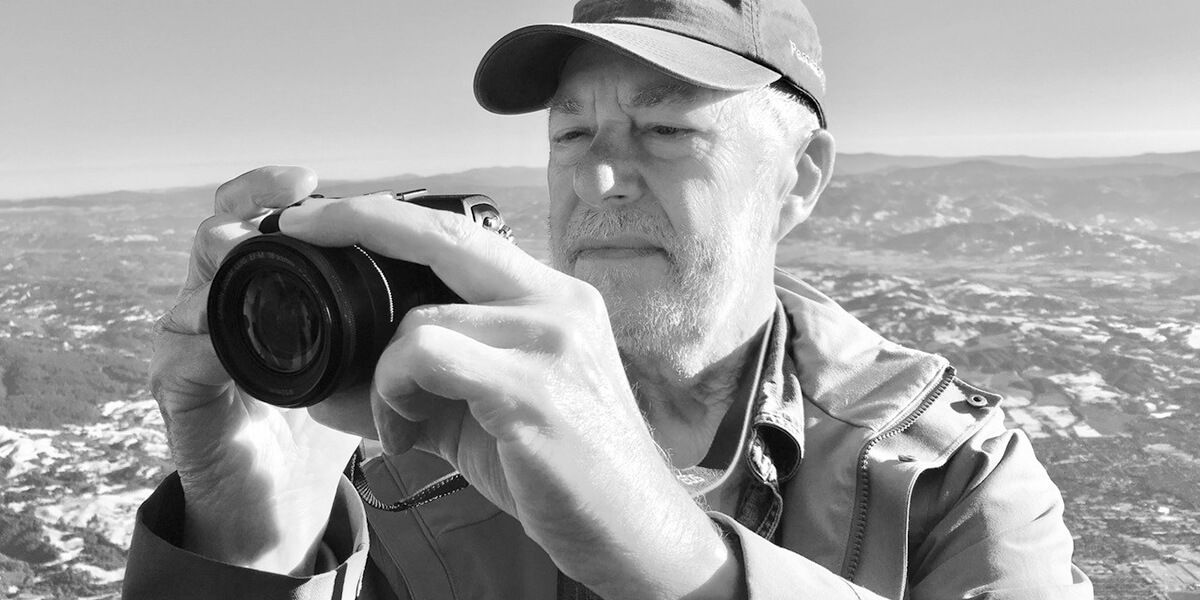 Roy Ramsing looking at a viewfinder of a camera while standing on the summit of a mountain