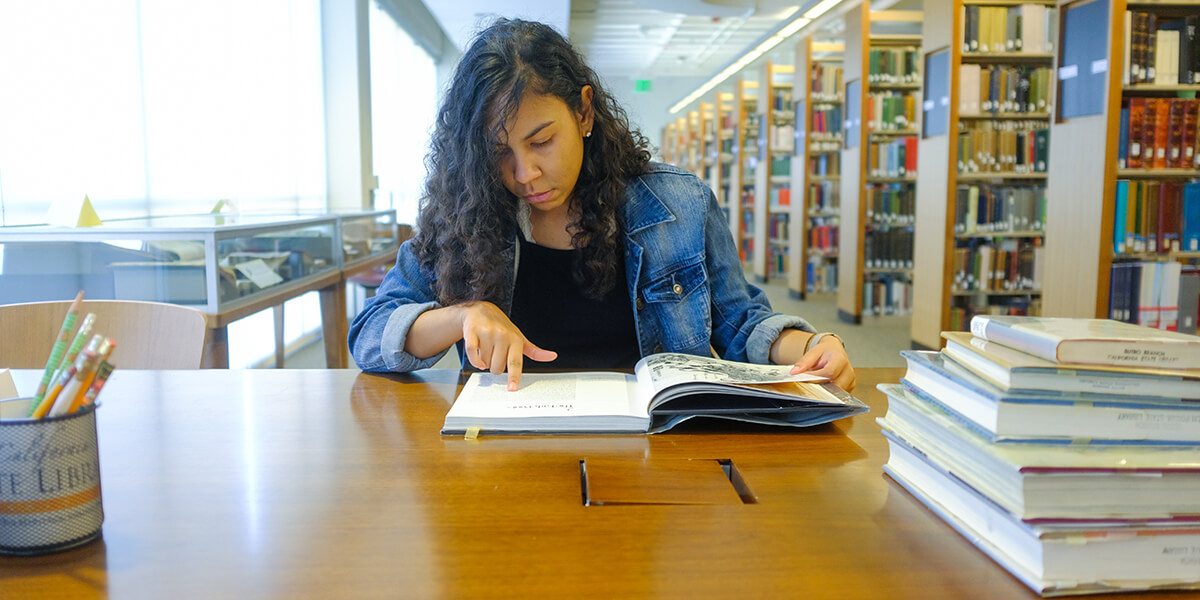 Photo of Kayla Ratliff pointing her finger on a page of a book while seated at a table in J. Paul Leonard Library