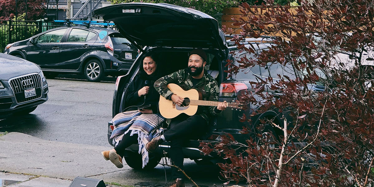 Photo of Rachel Garcia and Thu Tran perform acoustic guitar and vocals while seated in the hatchback of a car