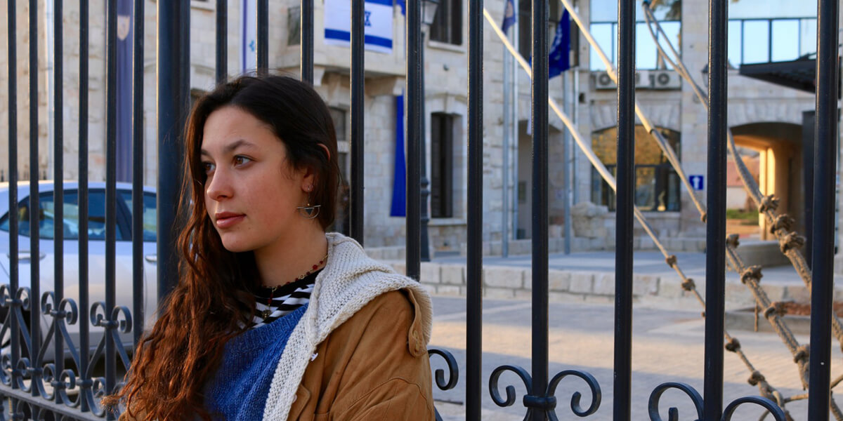 Photo of Atalya Ben-Abba leaning against fence, with view of building with Israel flag in background