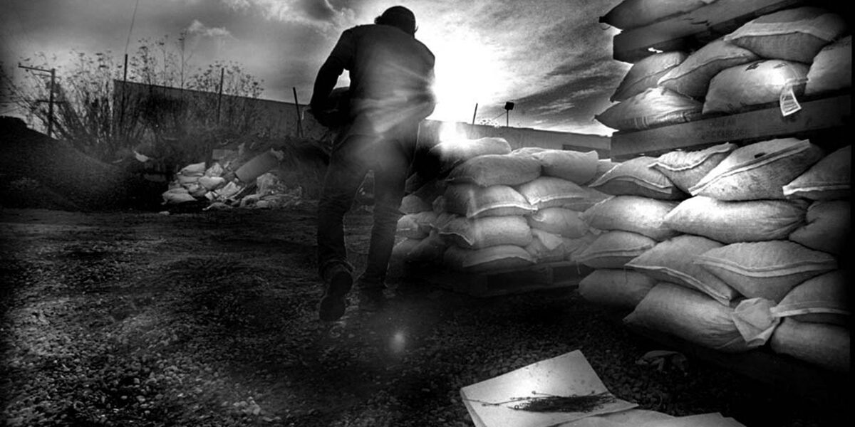Black and white photo of man carrying sandbag next to piles of sandbags with the sun shining in the distance