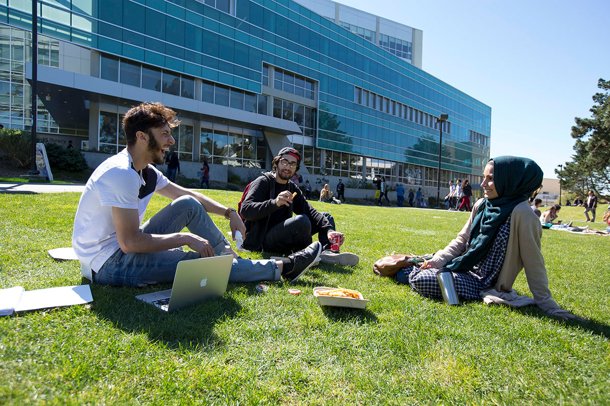 Three students sit on lawn with J Paul Library in background