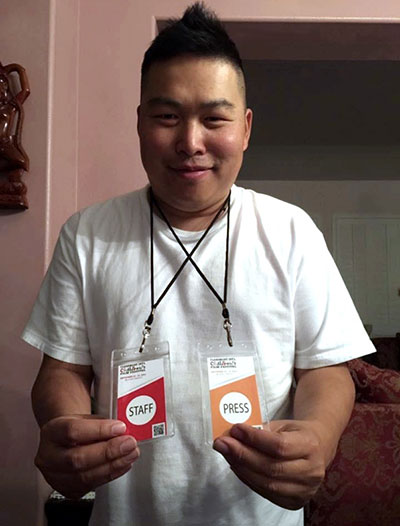 Man wearing a white T-shirt smiles at the camera while holding two event badges labeled “Staff” and “Press” in an indoor setting.