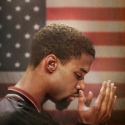 Mahmood Abdul-Rauf holds his hands in prayer in front of the USA flag while wearing a Denver Nuggets warmup jacket