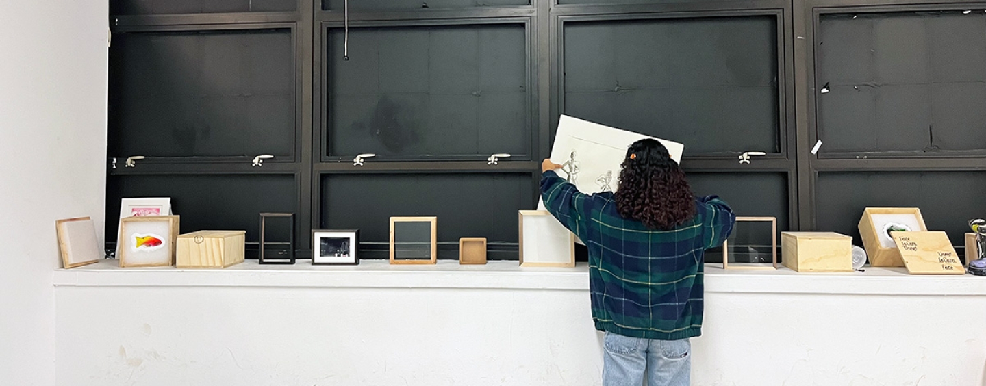 A student is standing in a room, facing away from the camera, organizing or examining items on a counter beneath large wall-mounted cabinets. The cabinets are open, displaying empty shelves. Atop the counter are various books and small boxed items.