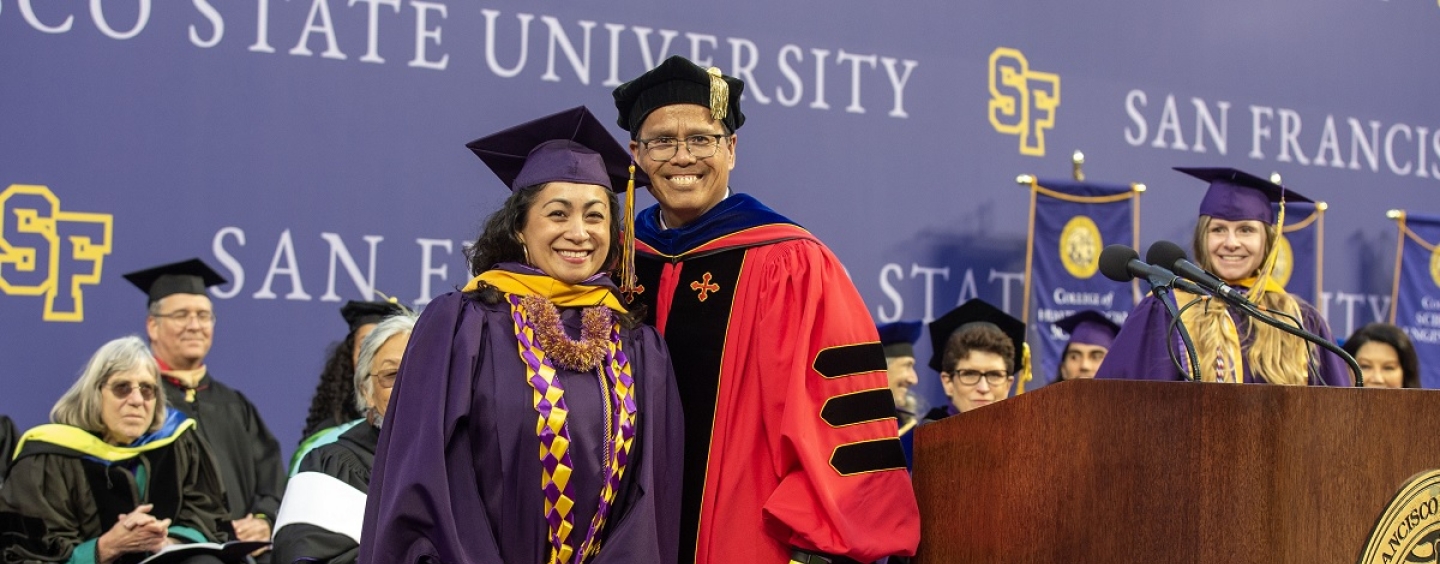 A woman and man in Commencement regalia smile at the camera