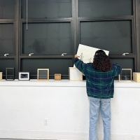 A student is standing in a room, facing away from the camera, organizing or examining items on a counter beneath large wall-mounted cabinets. The cabinets are open, displaying empty shelves. Atop the counter are various books and small boxed items.