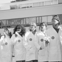 Black and white photo of eight SFSU students in lab coats standing on the Quad with four of them each eating a slice of pizza