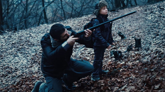 A man points a rifle in the woods with a child standing next to him.
